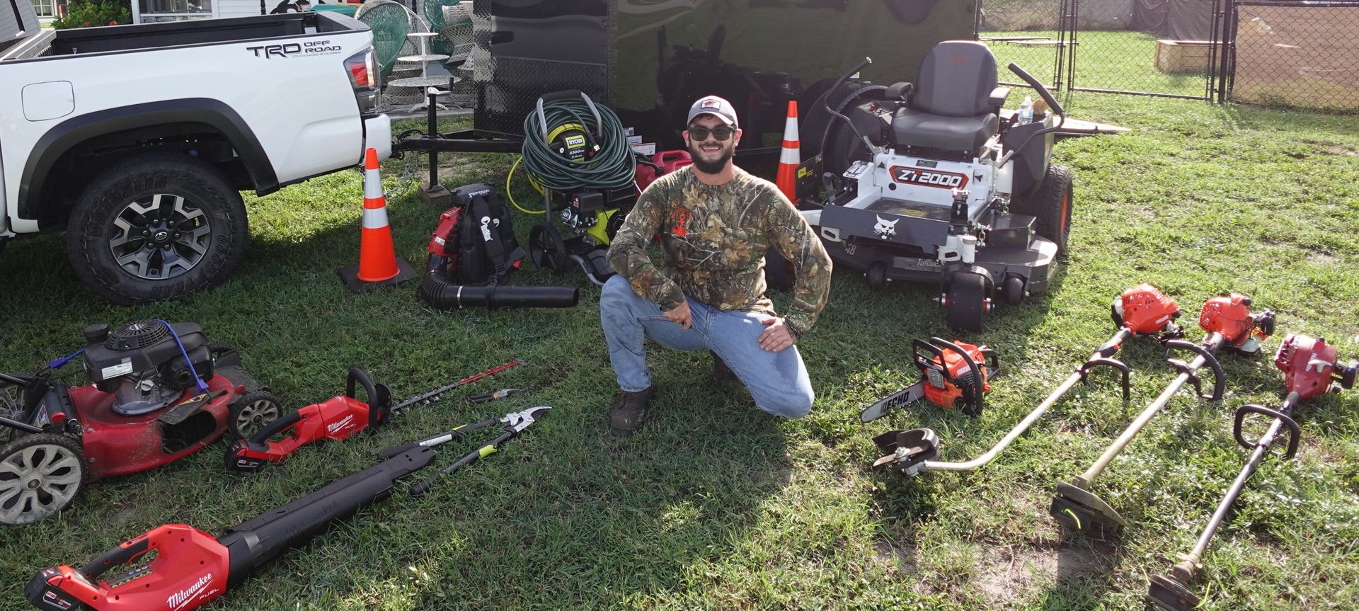 Collin kneeling confidently and smiling in front of his truck, trailer, mower, and full lawn care equipment lineup
